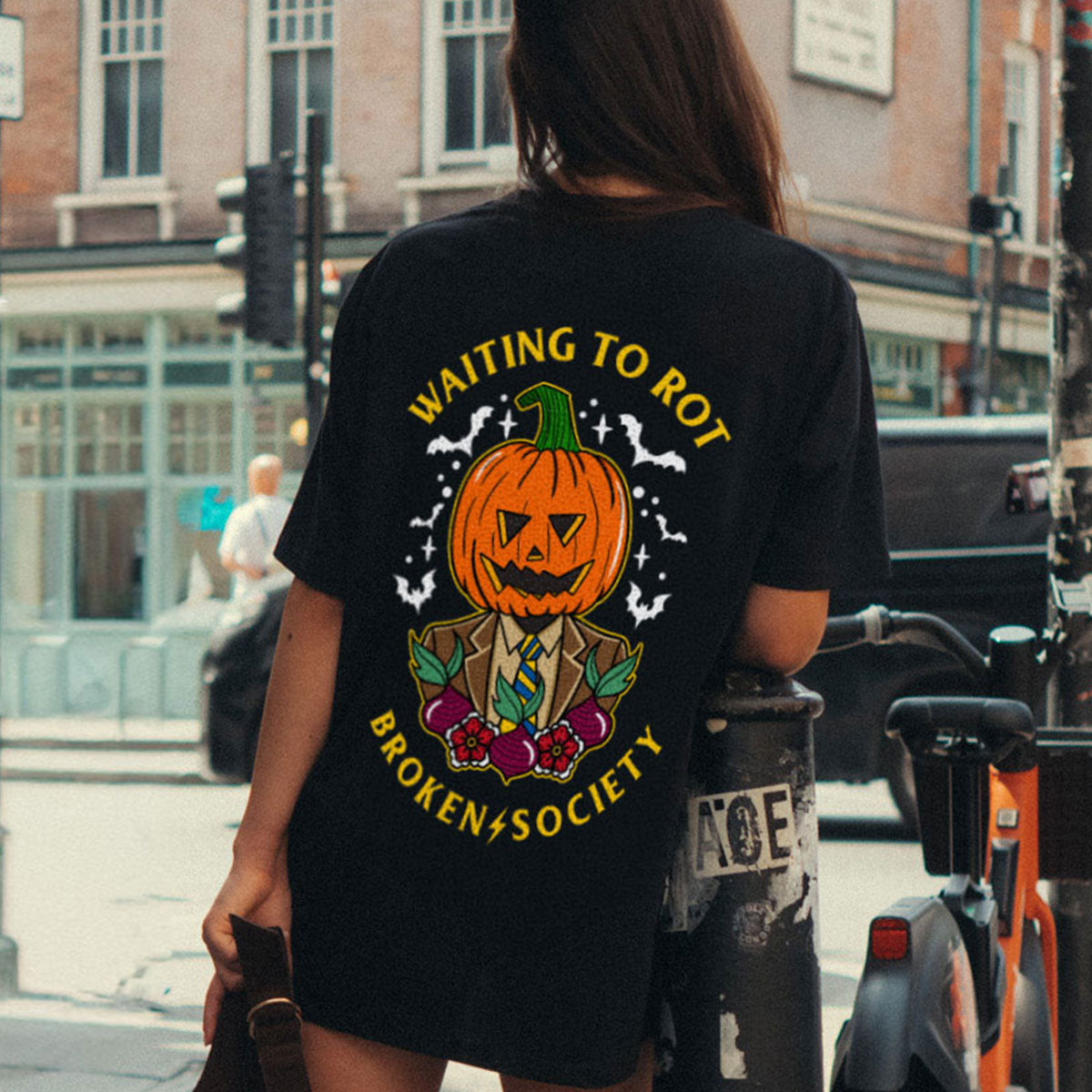 A woman viewed from behind wearing a black graphic t-shirt with a Halloween pumpkin design and the text 'WAITING TO ROT' and 'BROKEN+SOCIETY'.