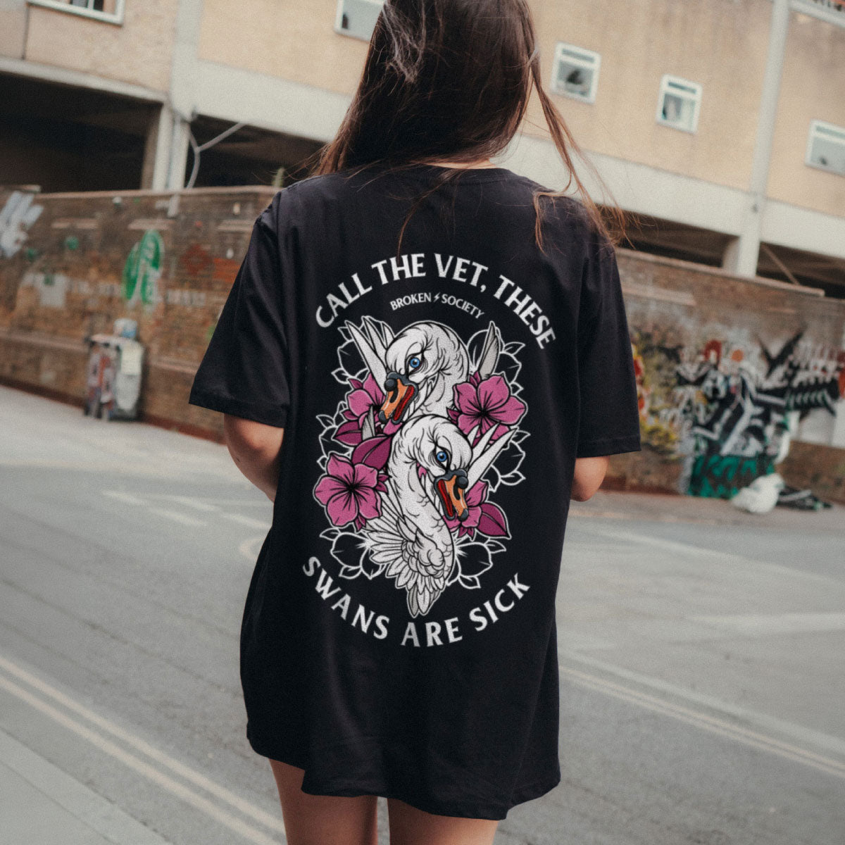 A woman viewed from behind wearing a black oversized T-shirt with a large graphic print of two white swans and pink flowers, with the text 'CALL THE VET, THESE SWANS ARE SICK' on the back, in an outdoor urban setting.