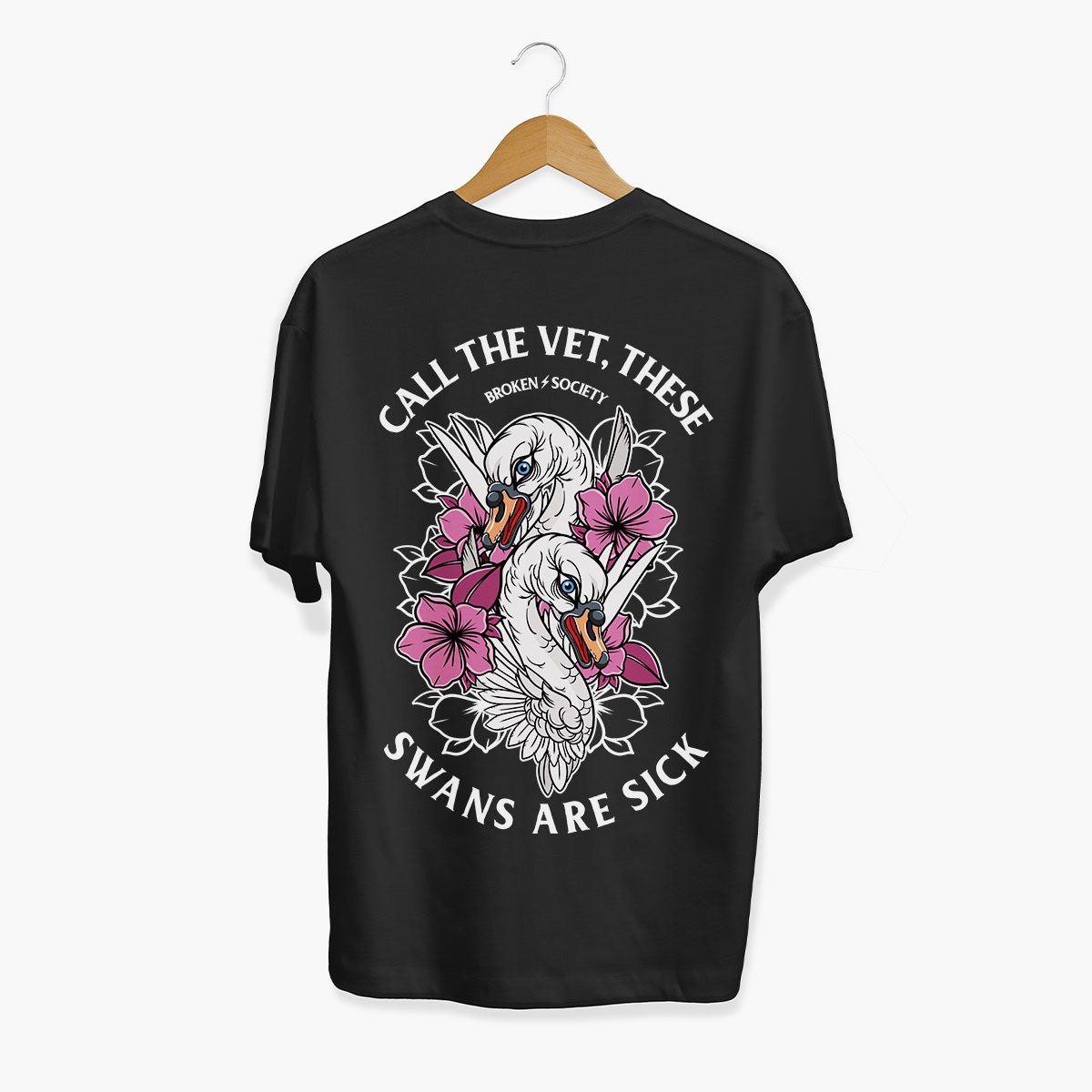 A black short-sleeve t-shirt hanging on a wooden hanger against a light gray background. The back of the shirt features a large graphic print of two white swans surrounded by pink flowers, with the text 'CALL THE VET, THESE SWANS ARE SICK' and 'BROKEN SOCIETY' written in white.