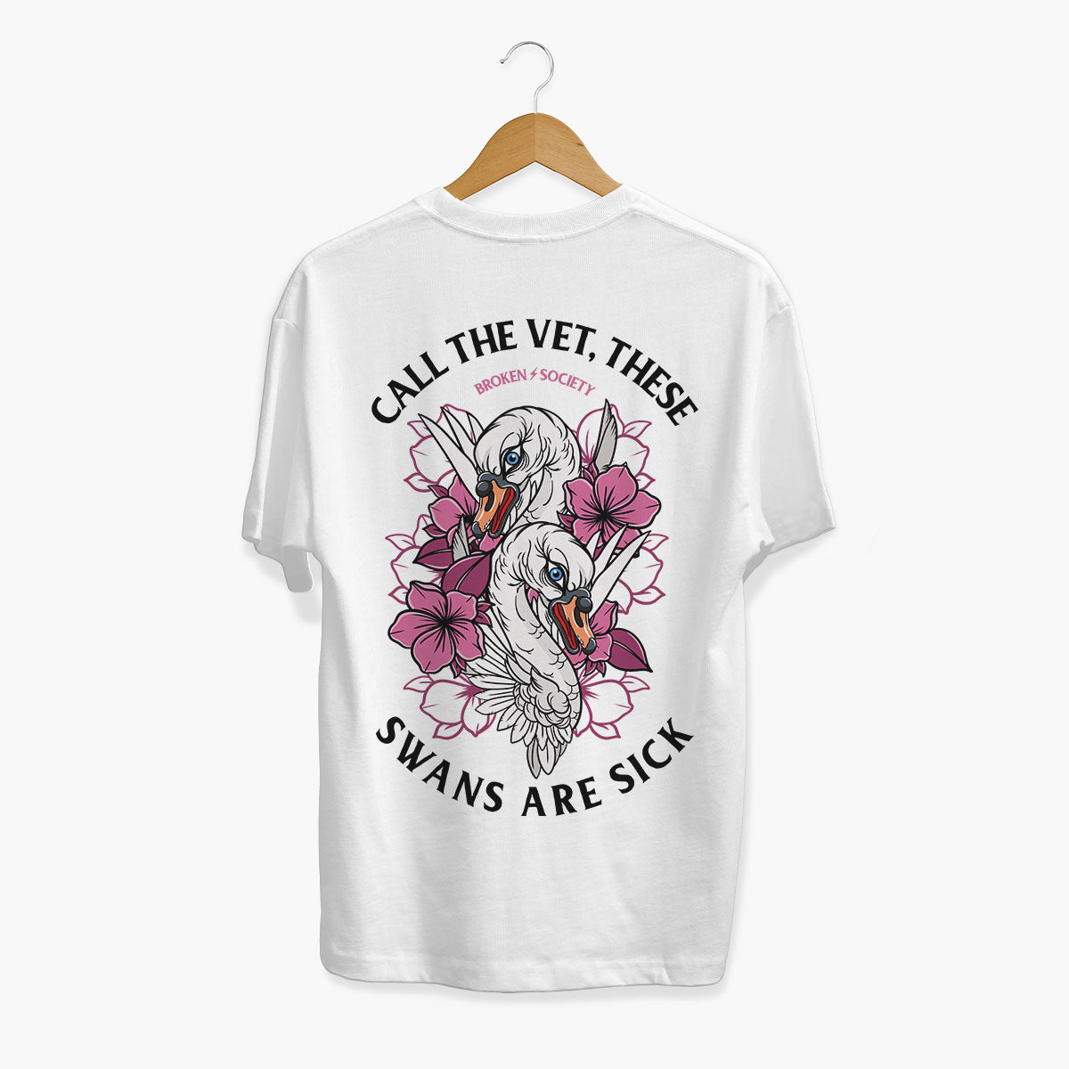 A white short-sleeve t-shirt hanging on a wooden hanger against a light gray background. The back of the shirt features a large graphic print of two swans surrounded by pink flowers, with the text 'CALL THE VET, THESE SWANS ARE SICK' and 'BROKEN SOCIETY' printed in black and pink.